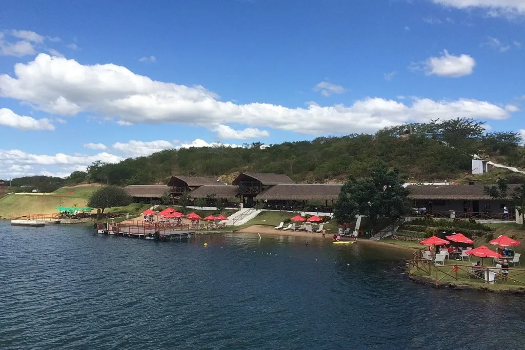Vista de um resort à beira de um lago com espreguiçadeiras, guarda-sóis vermelhos e área de praia, em meio a colinas verdes, sob um céu azul com nuvens brancas.