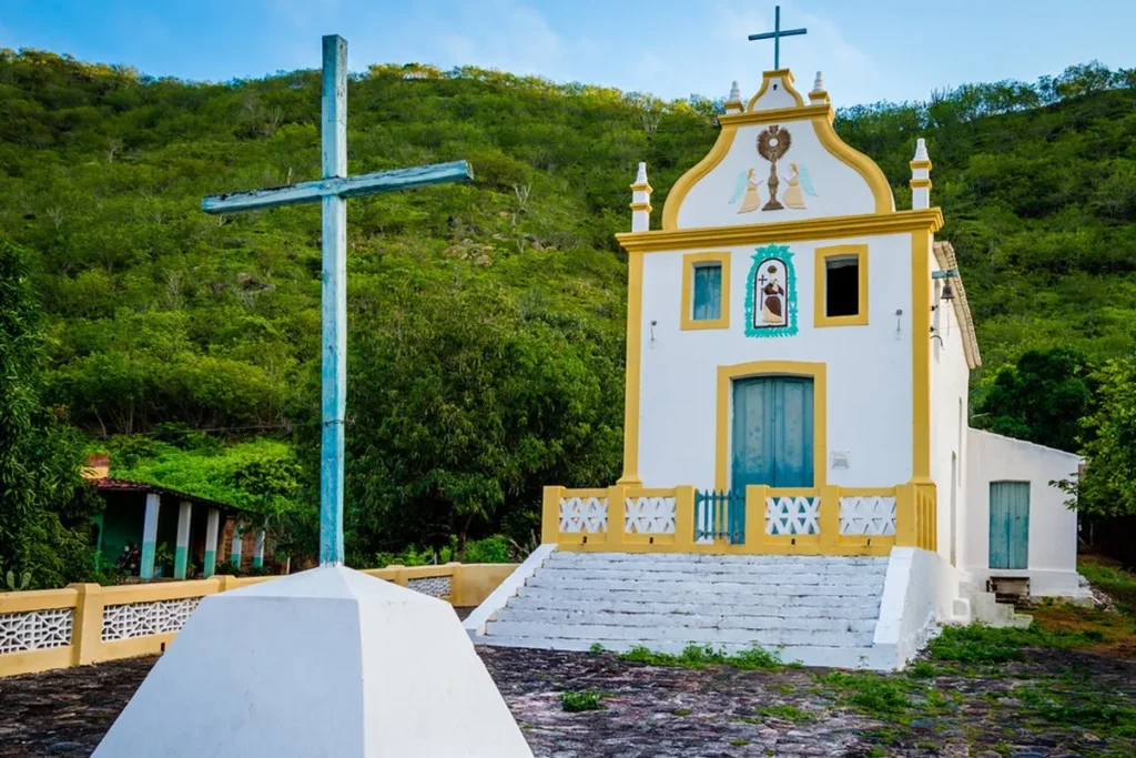 Igreja de Santo Antônio de Lisboa com fachada branca e detalhes em amarelo, cruz em primeiro plano, cercada por vegetação